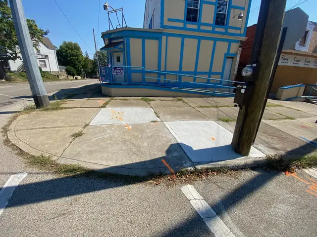 Street-corner building with pale siding and bright blue trim; patched sidewalk and utility markings in the foreground.