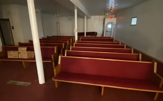 Interior hall lined with wooden pews with burgundy cushions, chandeliers, and white columns.