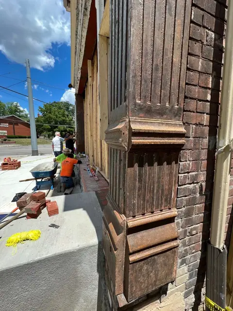 Masonry crew beside a red brick wall next to a rusted ornate cast-iron column; brick stacks and levels on the ledge.