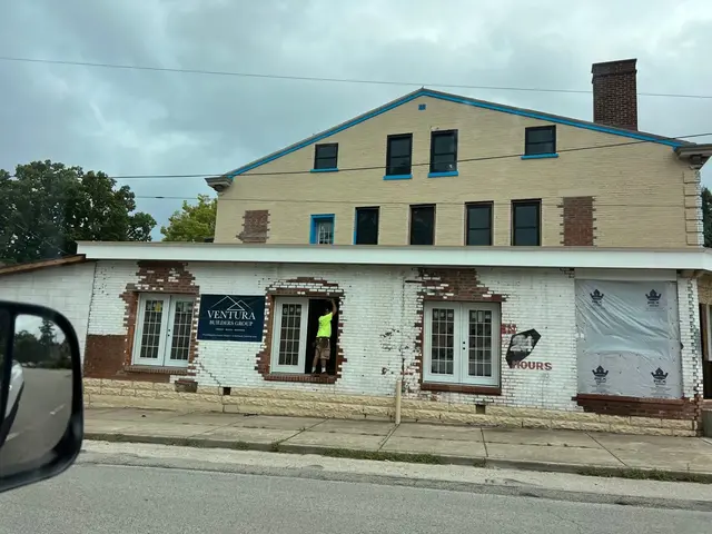 Worker in neon shirt stands in a new doorway; Ventura Builders Group banner on white brick that shows patches of red underneath.
