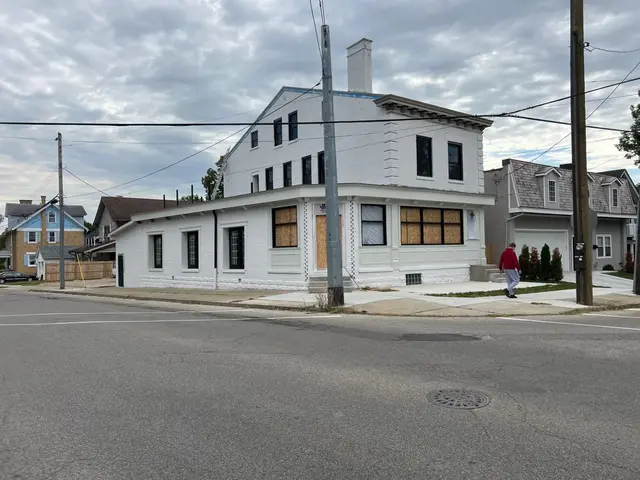 White-painted brick corner building with multiple ground-floor openings boarded in plywood; pedestrian on the sidewalk.