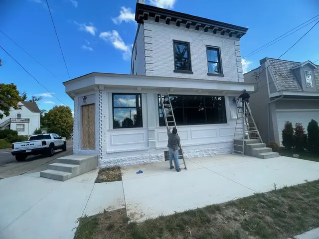 Two painters on ladders working white trim above black-framed windows on the refreshed façade.