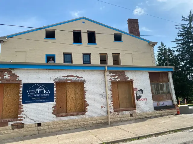 Front elevation with tan upper brick and white lower brick; Ventura Builders Group banner and several boarded openings.