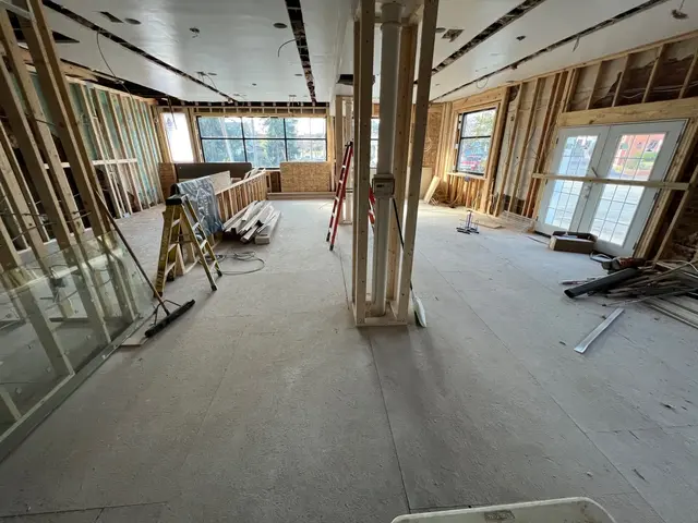 Large gutted room with exposed studs, partially finished ceiling panels, ladders, and dust-covered subfloor.