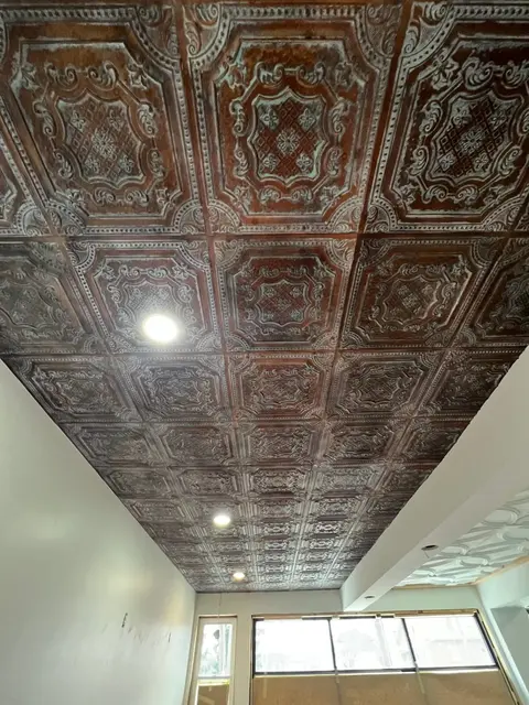 Ornate pressed-metal ceiling tiles with copper tone and recessed lights against freshly painted white walls.