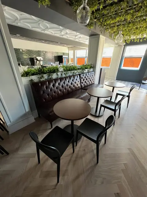 Tufted leather banquette, round wood café tables, black chairs, herringbone flooring, and green tiled wall beyond.