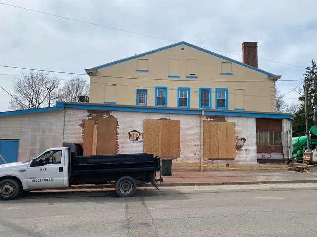 Two-story tan and white brick building with blue trim; plywood-covered openings and a dump truck parked along the curb.