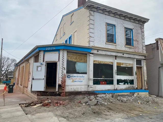 Corner view of former grocery with faded Self Service Market lettering; missing brick at base and debris piled on the sidewalk.